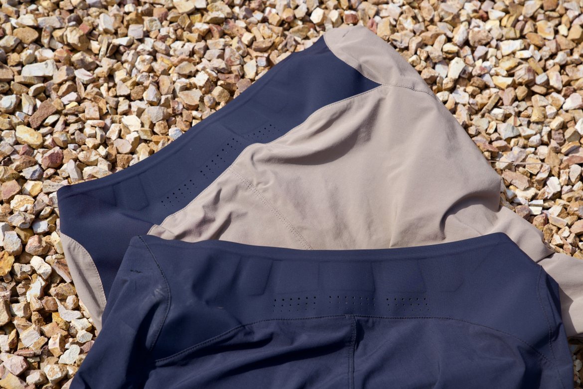 A close-up view of two pairs of sports shorts lying on a bed of small, multicolored gravel. One pair is navy blue and the other is a light beige, showing smooth fabric with a modern cut and subtle design details.
