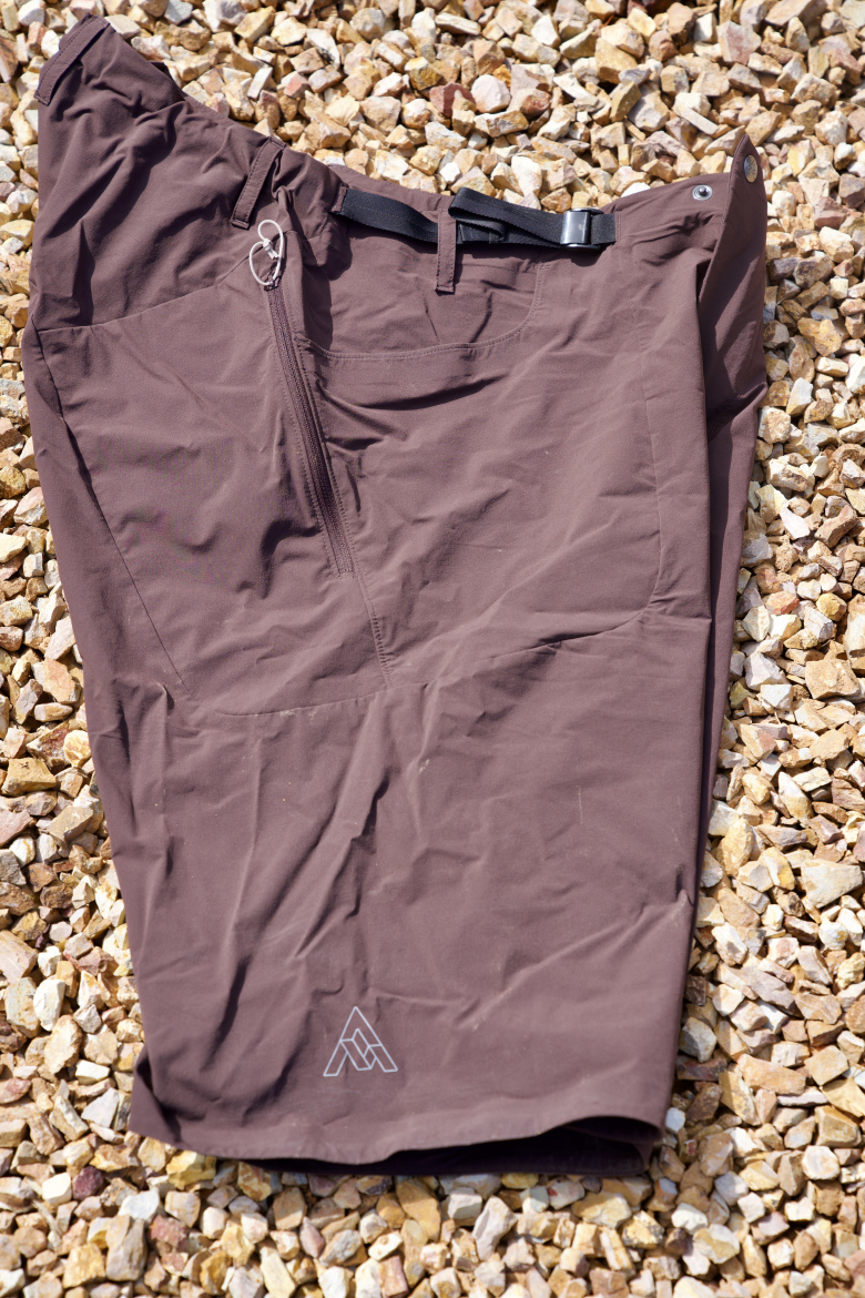 A pair of brown outdoor shorts laid flat on a bed of small, light-colored stones. The shorts feature a zipper pocket, a belt loop, and a logo on the lower left side. The fabric appears lightweight and is suitable for outdoor activities.