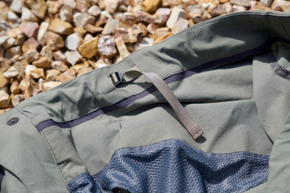 Close-up view of the interior of a green jacket featuring a mesh lining, adjustable straps, and a button closure, set against a background of small, varied stones.
