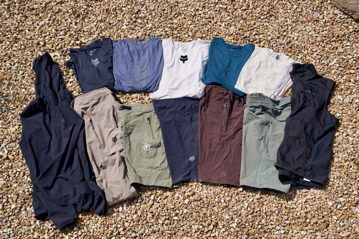 A variety of outdoor clothing laid out on a gravel surface, including t-shirts in shades of blue, gray, and white, as well as shorts and lightweight pants in earthy tones like beige, green, and brown, with some items featuring zippers and logo designs.