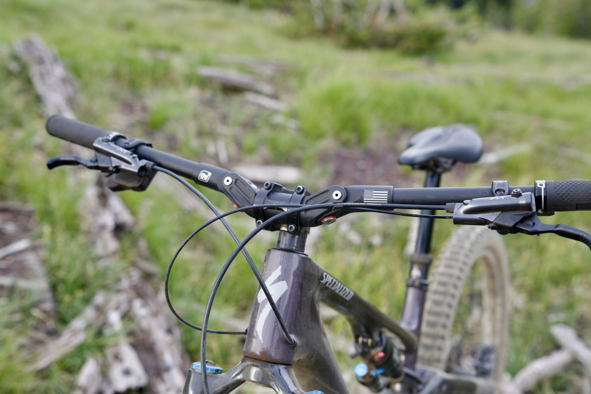 Close-up view of a mountain bike handlebar and frame, featuring a black grip, brake lever, and visible cables. The bike is positioned on a grassy area with some logs in the background, suggesting an outdoor cycling environment. The bike frame is branded "Specialized," and there's a small American flag decal on the handlebar.