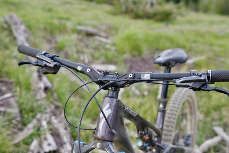 Close-up view of a mountain bike handlebar and frame, featuring a black grip, brake lever, and visible cables. The bike is positioned on a grassy area with some logs in the background, suggesting an outdoor cycling environment. The bike frame is branded "Specialized," and there's a small American flag decal on the handlebar.