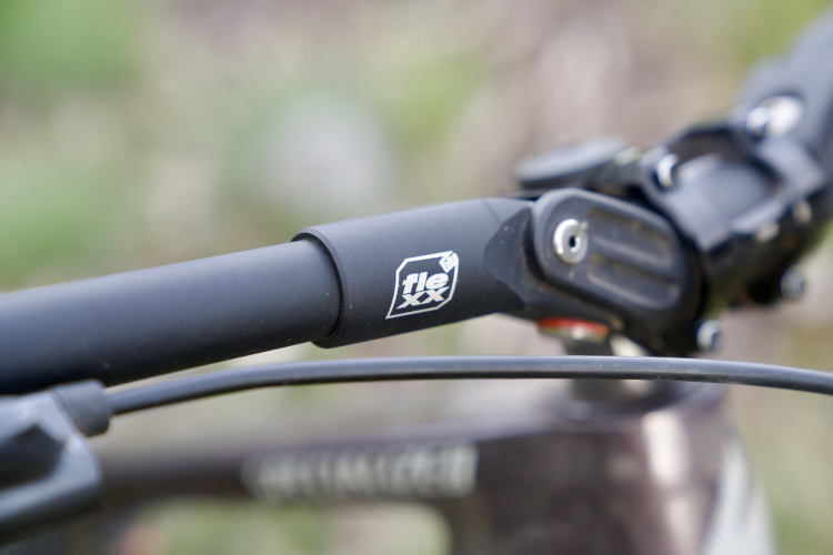 Close-up image of a bicycle's handlebar featuring a black stem with a label reading "flexxx." The handlebars are positioned against a blurred outdoor background with greenery, capturing details of the bike's components.