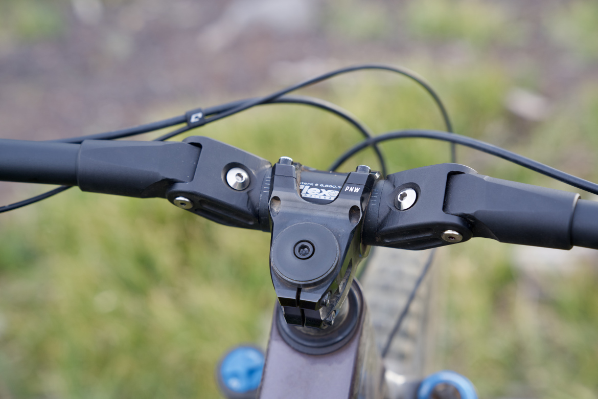Close-up view of a mountain bike handlebar, showing the stem, grips, and cables against a blurred green background.