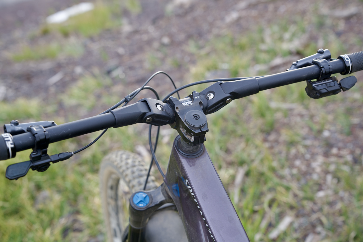 Close-up view of mountain bike handlebars, featuring a sturdy design with integrated brake and gear levers, a stem connector, and a control for adjusting a suspension system. The scene is set against a backdrop of green grass and earthy terrain, suggesting an outdoor biking environment.