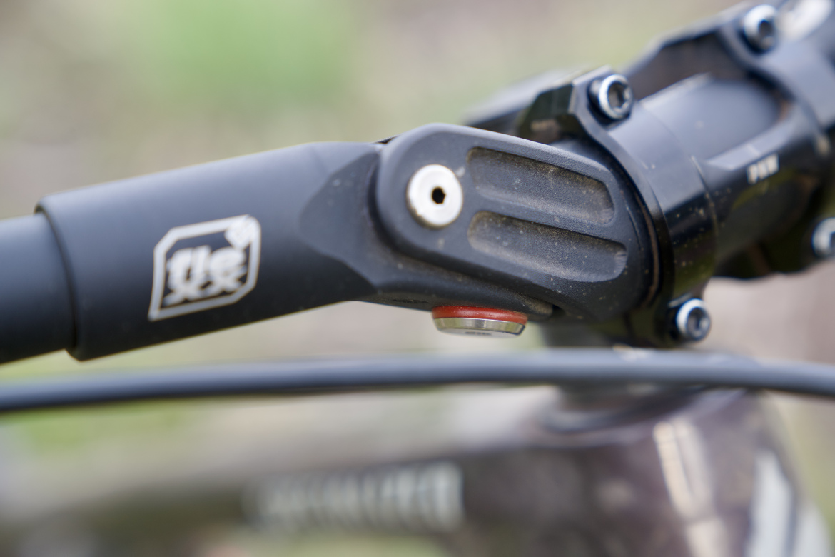 Close-up image of a bike handlebar featuring a black stem with visible branding and a red adjustment button. The handlebar shows signs of usage, with some dirt and scratches, set against a blurred natural background.