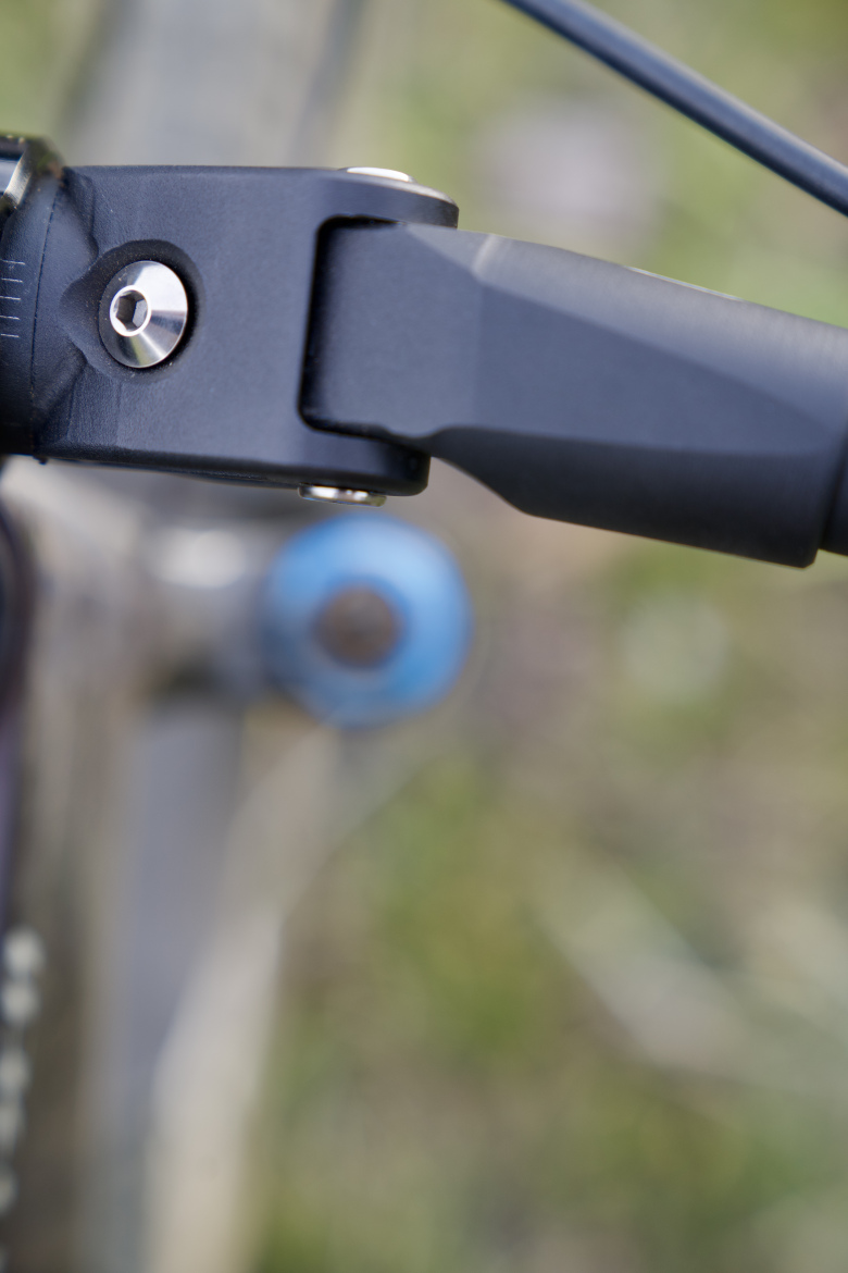 Close-up image of a bicycle handlebar component, showcasing a black lever and connecting hardware against a blurred green background. The focus is on the detailed mechanical features, highlighting the craftsmanship.