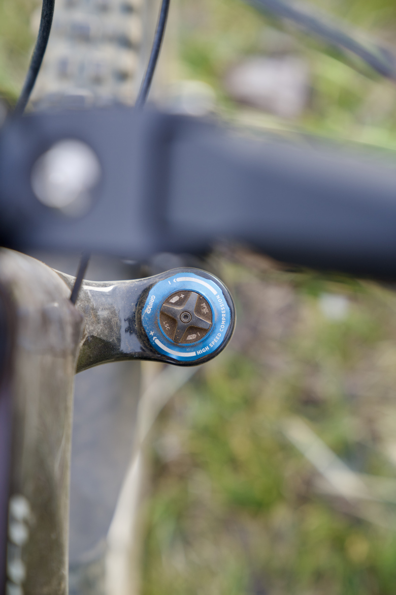 Close-up image of a bike's suspension adjustment knob, featuring a blue outer ring and a textured inner dial, set against a blurred grassy background. The knob is labeled "HIGH SPEED COMPRESSION."