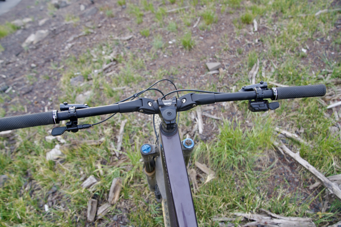 Close-up view of a mountain bike handlebar from a top-down perspective, featuring black grips, various control levers, and a sleek, dark frame. The background includes patches of grass and small twigs, suggesting an outdoor setting.