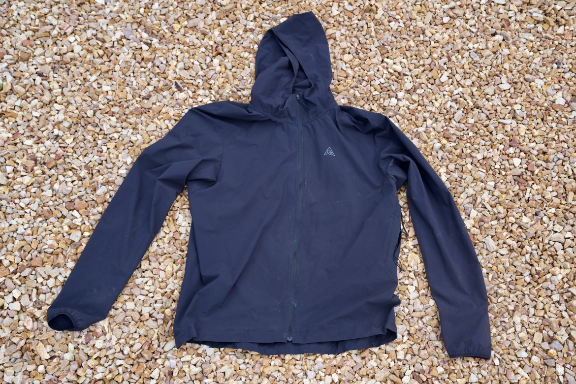 A black lightweight jacket with a hood laid flat on a surface covered with small pebbles. The jacket features a full front zipper and has a logo on the upper left side.
