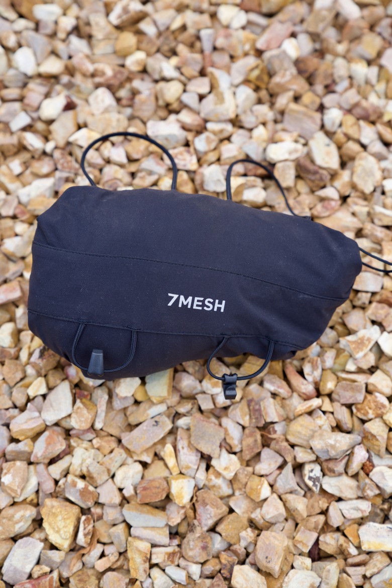 A black outdoor bag with the brand name "7MESH" displayed on the side, resting on a bed of small, multicolored gravel stones. The bag features multiple straps for securing or carrying.