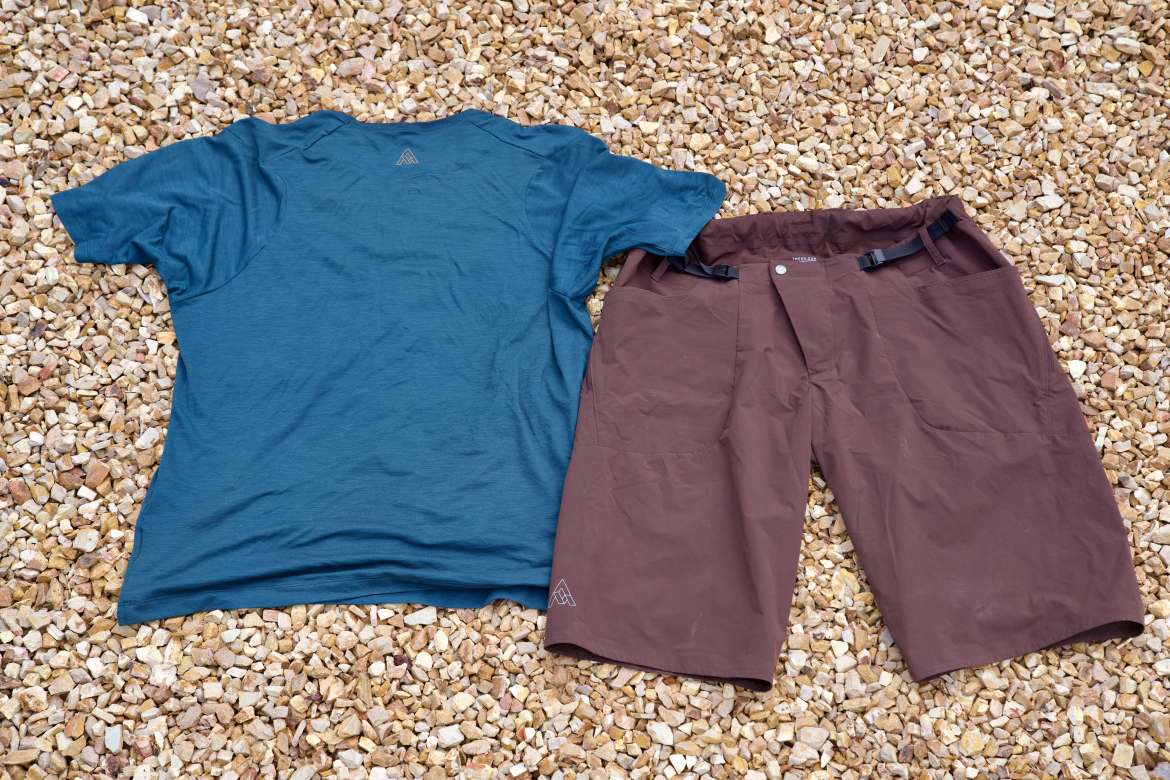 A blue short-sleeve shirt and brown shorts are laid out flat on a bed of small, multicolored gravel stones. The shirt is positioned on the left, with the neckline and shoulders visible, while the shorts are on the right, featuring a button and adjustable straps at the waist.