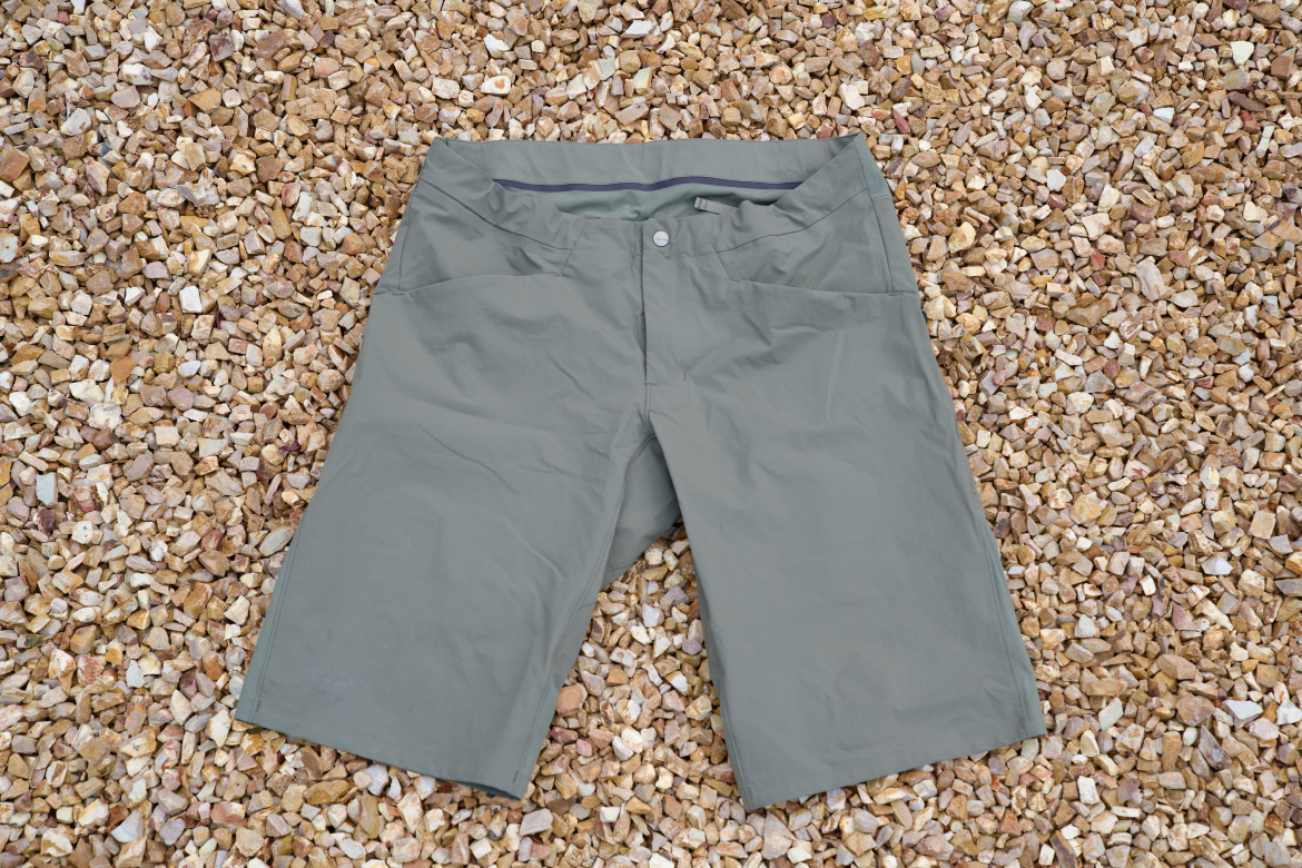 A pair of gray outdoor shorts laid flat on a surface covered with small, smooth pebbles. The shorts feature two front pockets and a button closure at the waist.