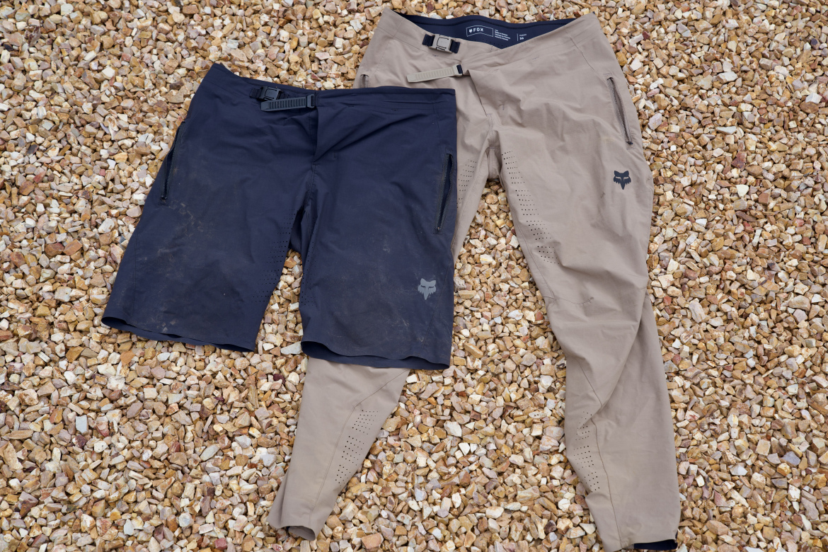 A pair of outdoor shorts, one in navy with dirt stains and a side zipper pocket, and the other in tan with a sleek design and ventilation details. The shorts are laid out on a surface covered in small pebbles.