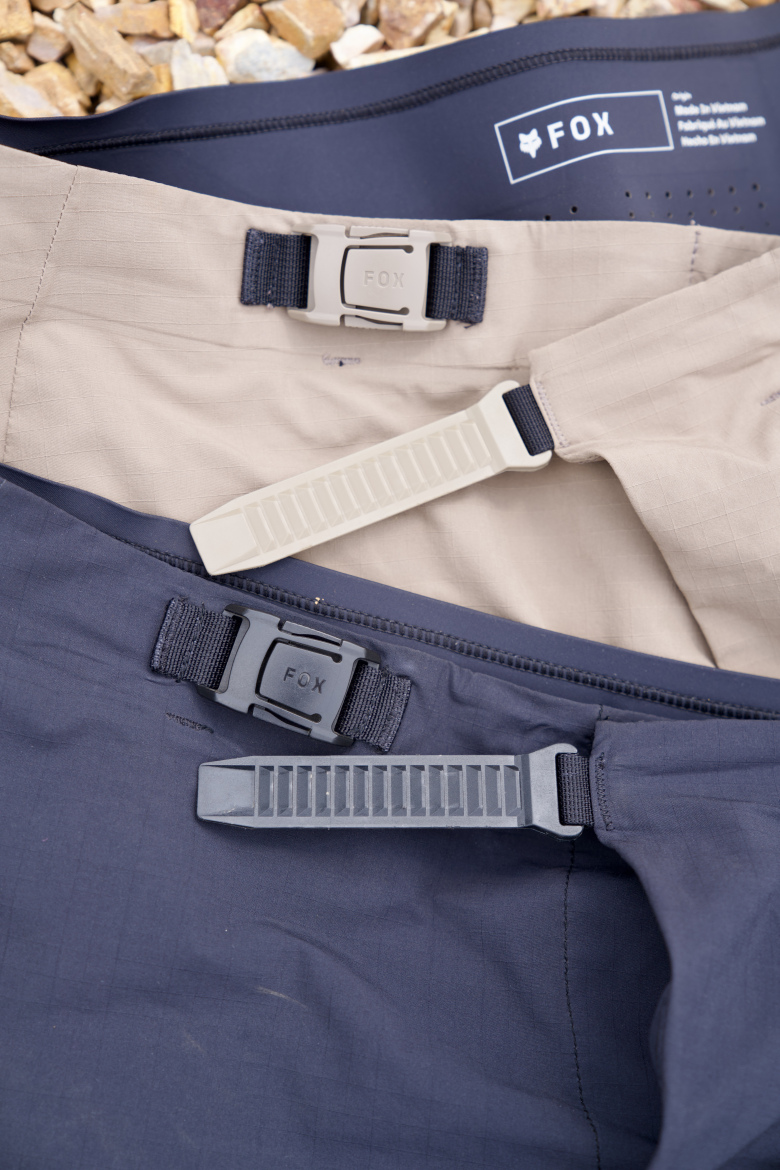 A close-up view of three pairs of shorts stacked on top of each other, showcasing different colors. Each pair features a distinctive strap and clasp design, with the brand name "FOX" visible on the clasps and tags. The background consists of small stones, creating an outdoor feel.