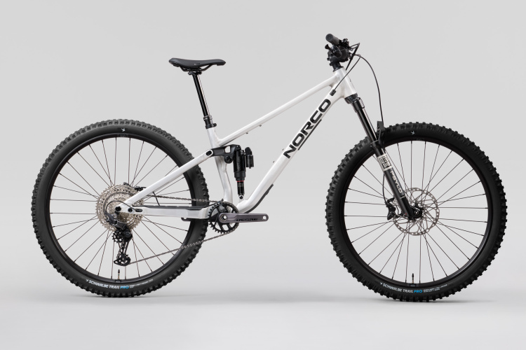 A white Norco mountain bike is displayed against a neutral gray background. The bike features wide, knobby tires, a robust frame, and a full suspension system. It is positioned to showcase its profile, with gear components and a disc brake clearly visible.