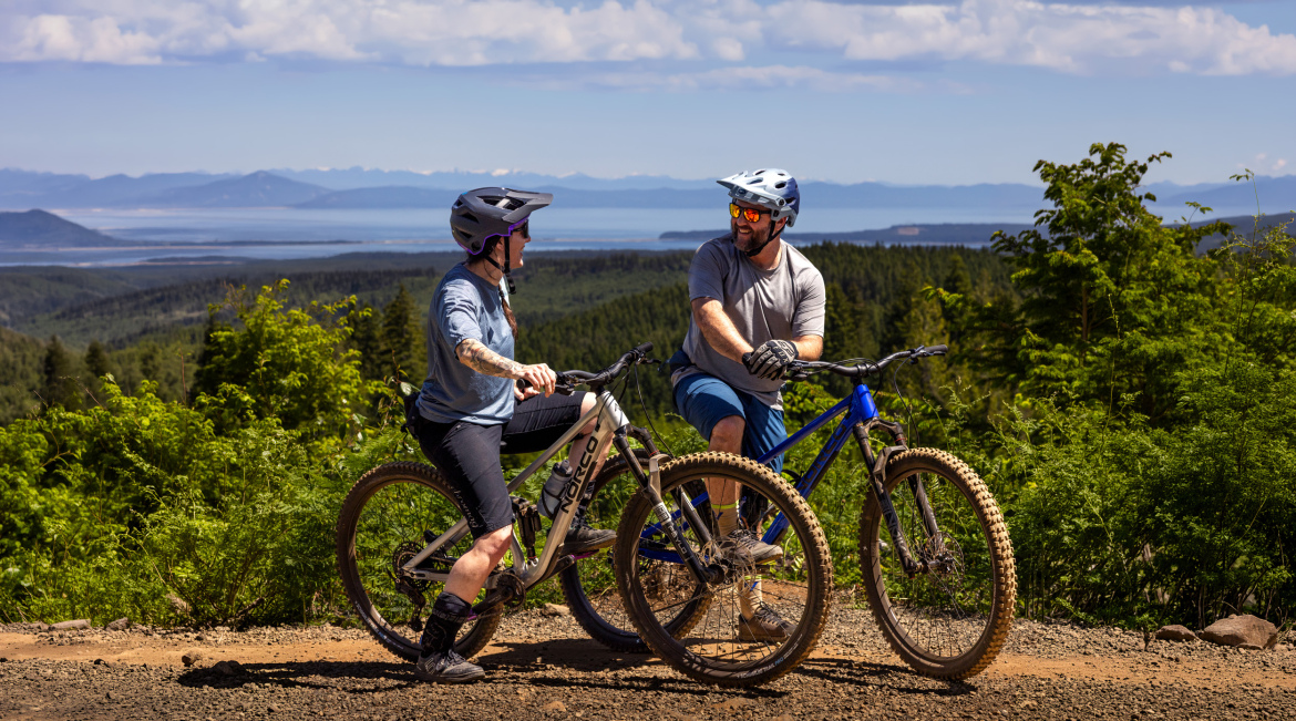 Two mountain bikers, one wearing a purple helmet and the other in a blue helmet, pause on a dirt trail surrounded by lush greenery and a scenic view of mountains and water in the background. They are engaged in conversation, both smiling and enjoying the sunny day. The rider on the left has visible tattoos and is dressed in a casual shirt and dark shorts, while the rider on the right is wearing a grey shirt and blue shorts.
