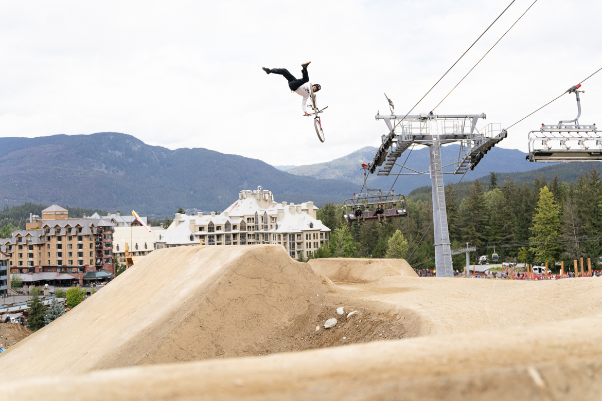 A BMX rider performing a mid-air trick above a dirt jump, with a scenic mountain backdrop and a ski lift in the foreground. Nearby, several buildings and trees are visible, indicating an outdoor recreational area. The atmosphere appears lively, likely during a biking event or competition.