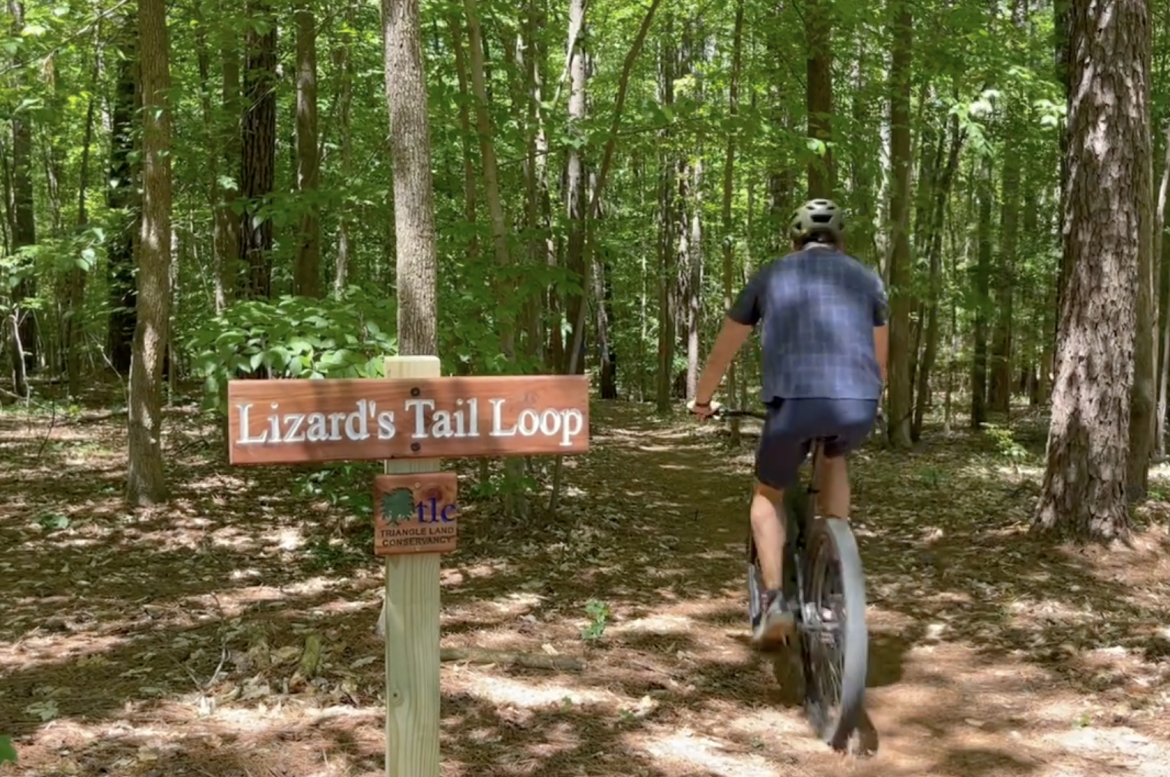 A cyclist riding a mountain bike on a forest trail near a wooden sign indicating "Lizard's Tail Loop," surrounded by lush green trees and natural scenery.