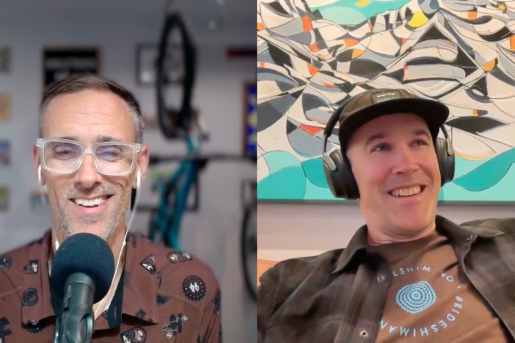 Two men are engaged in a conversation over a video call. The man on the left is wearing glasses and a brown patterned shirt, smiling and speaking into a microphone. The man on the right, wearing a black cap and headphones, is also smiling. A colorful abstract painting is visible in the background, along with a bicycle hanging on the wall behind the first man.