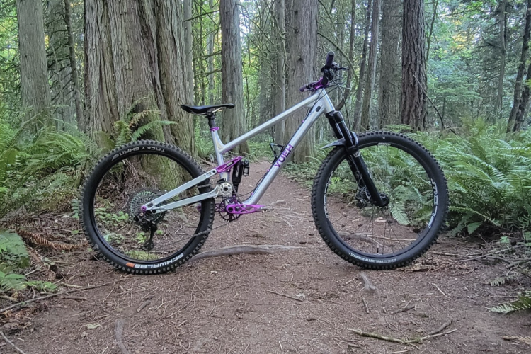 A mountain bike with a silver frame and purple accents is parked on a dirt trail surrounded by tall trees and ferns in a lush forest setting. The bike features thick tires and a front suspension fork, indicating it is designed for off-road use.