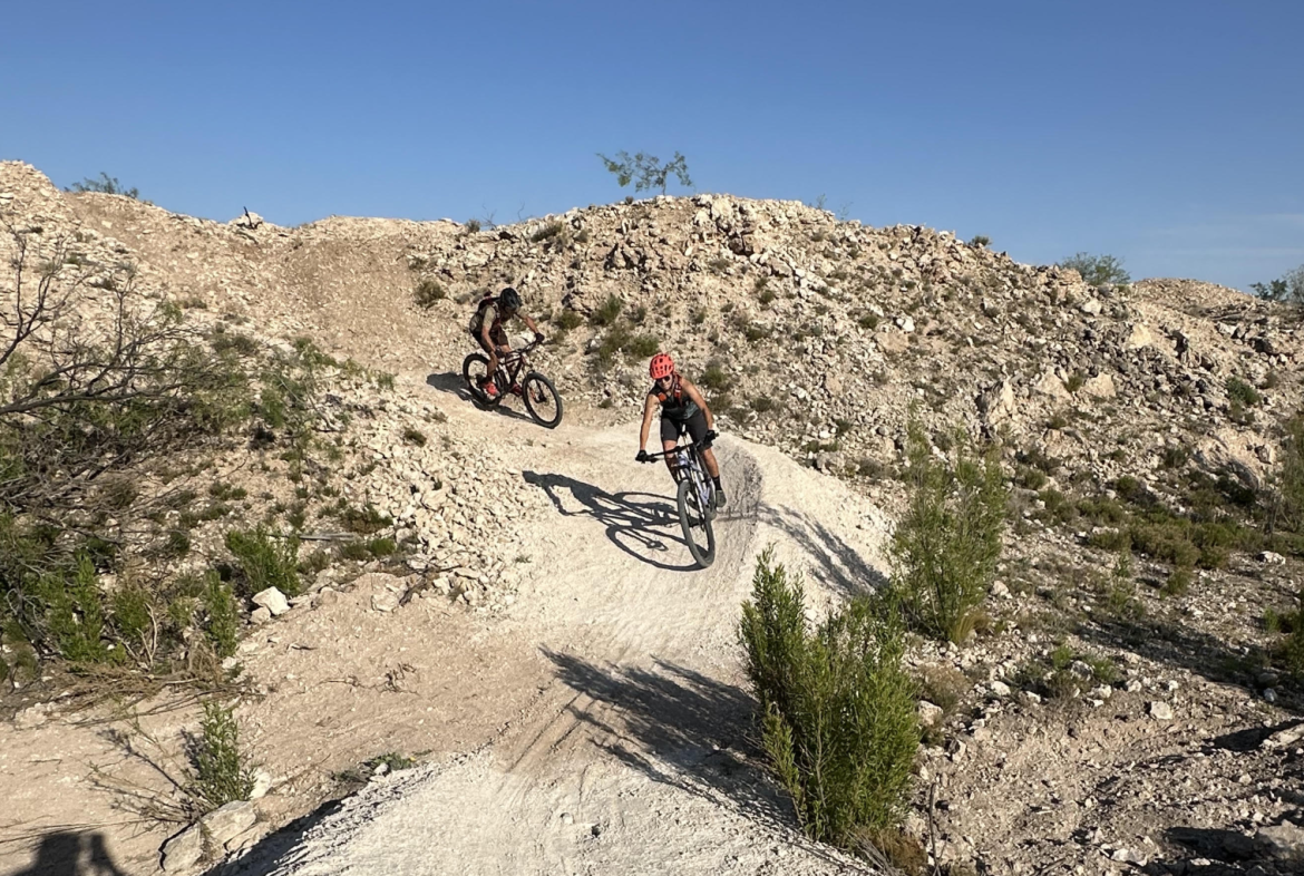Two mountain bikers navigating a dirt trail in a rocky terrain under a clear blue sky. One rider is in the foreground and appears to be leaning into a curve, while the other cyclist is further back on the trail. Sparse vegetation and rocky outcrops surround the biking path, creating a rugged outdoor setting.