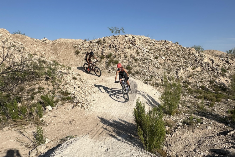 Two mountain bikers navigating a dirt trail in a rocky terrain under a clear blue sky. One rider is in the foreground and appears to be leaning into a curve, while the other cyclist is further back on the trail. Sparse vegetation and rocky outcrops surround the biking path, creating a rugged outdoor setting.