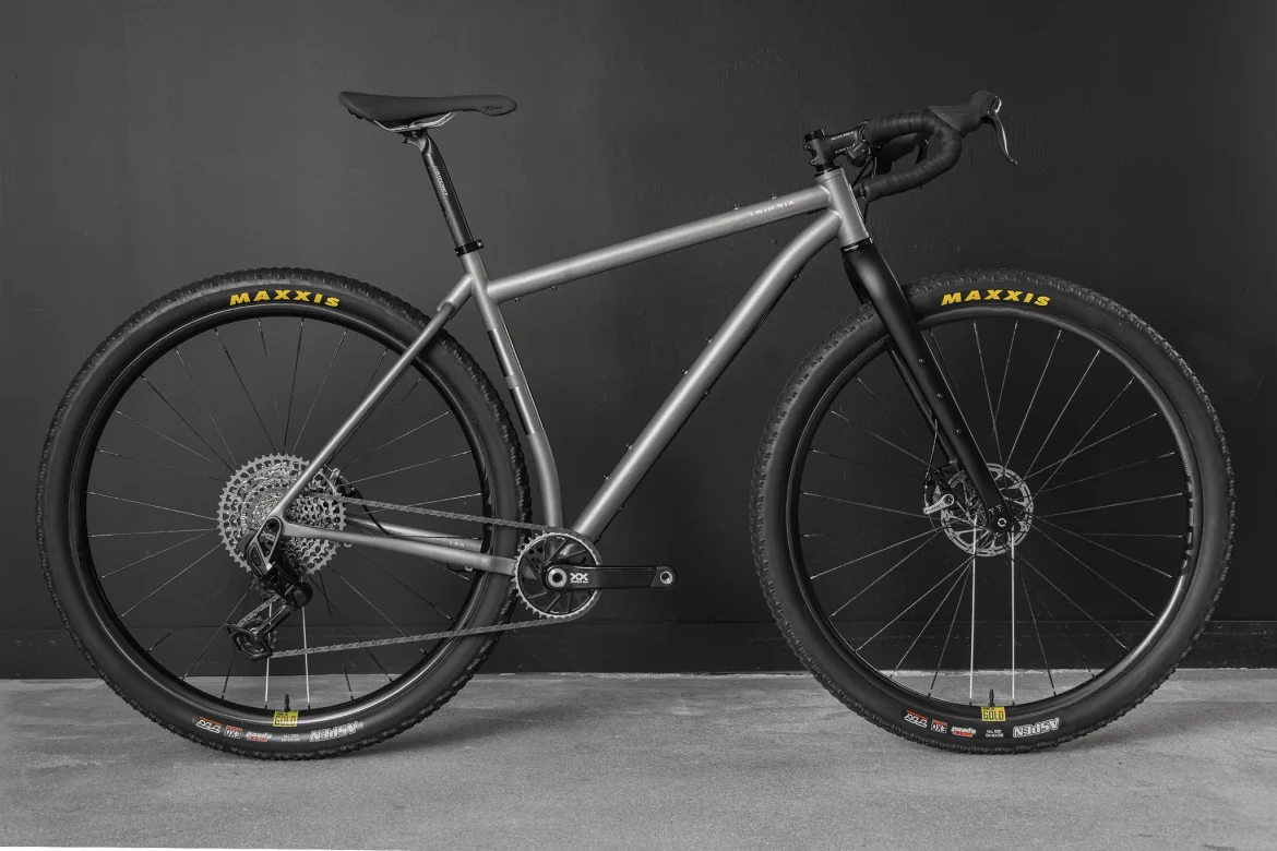 A modern bicycle with a sleek silver frame, featuring black wheels and Maxxis tires. The bike has a minimalist design, a single gear setup, and disc brakes. It is positioned against a dark wall, showcasing its streamlined structure and components.