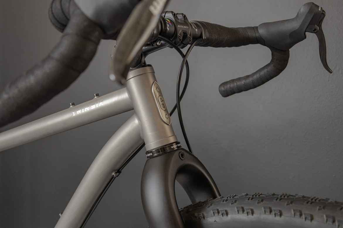 Close-up view of a gray bicycle frame featuring a sleek design, visible handlebars with black grips, and intricate detailing around the headset and fork area. The background is a simple dark gray, highlighting the bike’s components.