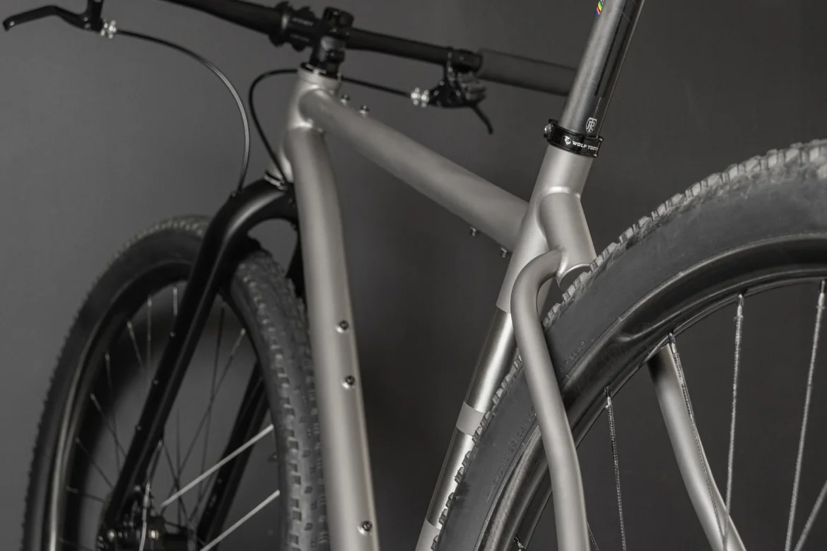 Close-up of a modern bicycle frame featuring a combination of matte black and silver finishes, showcasing the handlebars, fork, and tires against a dark background. The sturdy construction highlights the design details and tubing of the bike.
