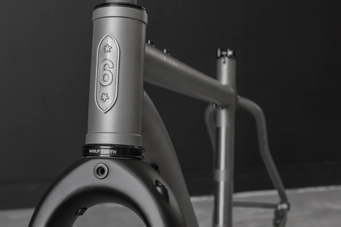 Close-up of a bicycle frame featuring a metallic finish, highlighting the number "6" and two stars on the head tube. The frame's design is sleek and modern, with a component bearing the name "WOLF TOOTH" adjacent to the fork. The background is a solid dark color, enhancing the bike's features.