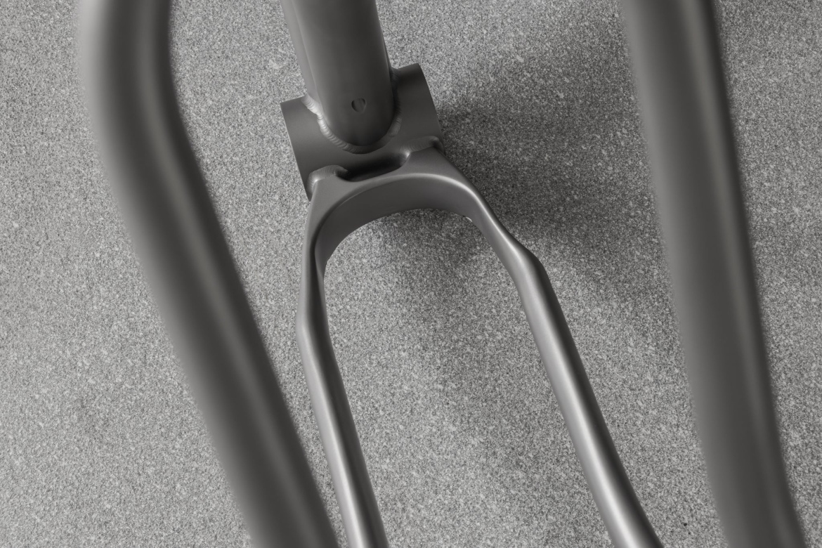 A close-up view of a metallic bicycle fork attached to a frame, showing the intricate design and welded joints, against a textured gray surface.