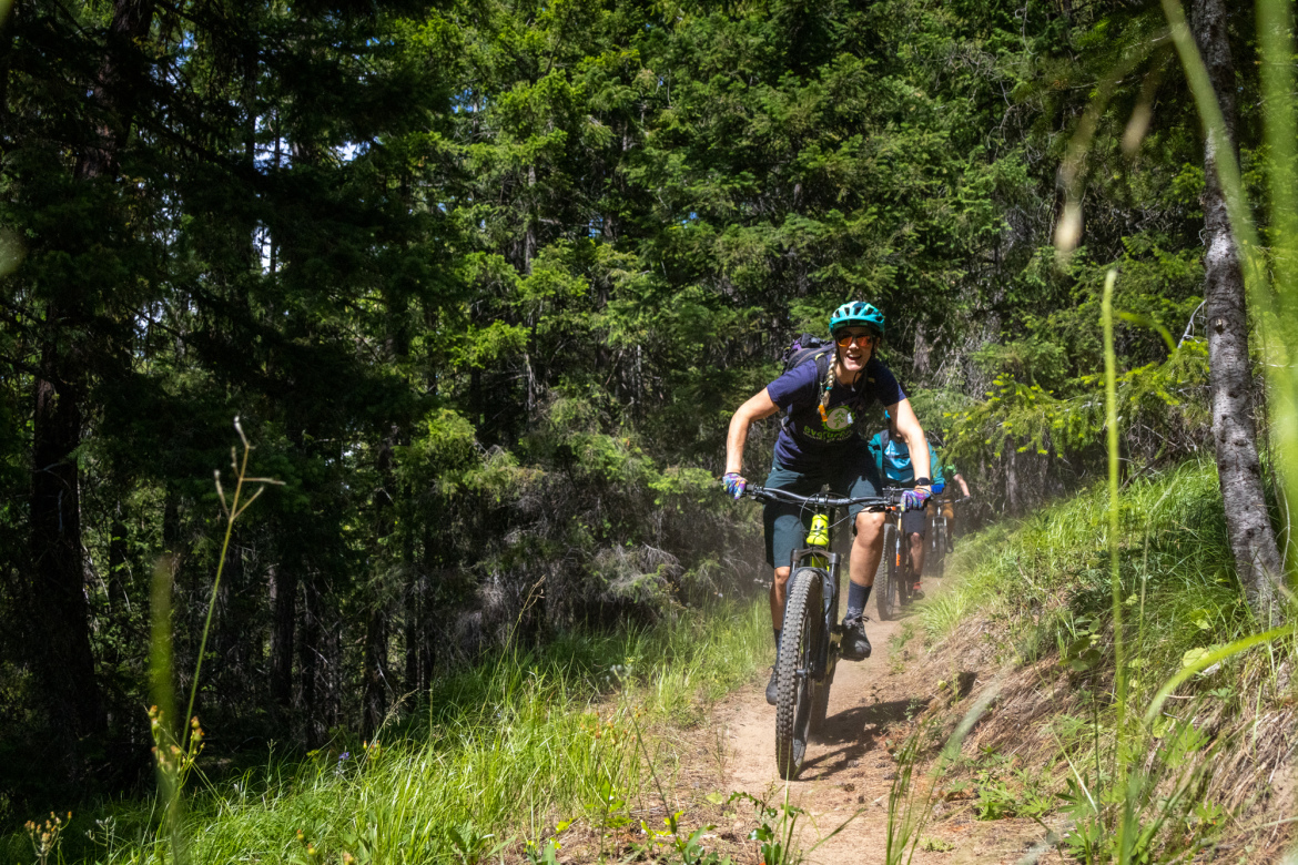 A group of mountain bikers riding down a dusty trail through a dense forest. The foreground shows a smiling rider wearing a blue helmet and a t-shirt, showcasing excitement as they navigate the path, with trees and green grass surrounding them. Another biker is visible in the background, adding to the sense of adventure in a natural setting.