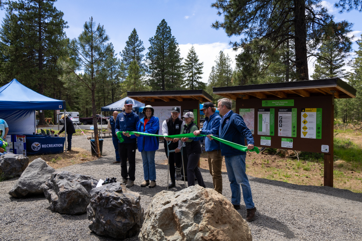 A group of six individuals stands in front of informational signs and tents at a recreational area, holding a large green ribbon. They appear to be participating in a ribbon-cutting ceremony, celebrating the opening of a new facility or trail. The background features trees and rocky landscaping, indicative of an outdoor setting.