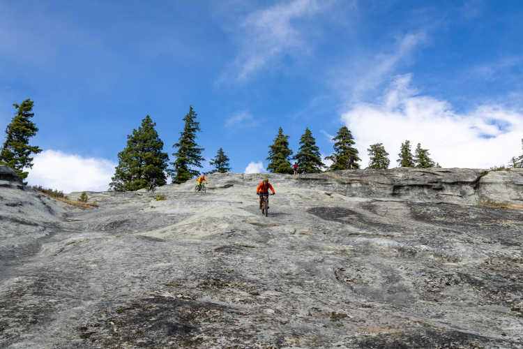 A group of mountain bikers navigating a rocky terrain under a blue sky with scattered clouds and tall pine trees in the background.