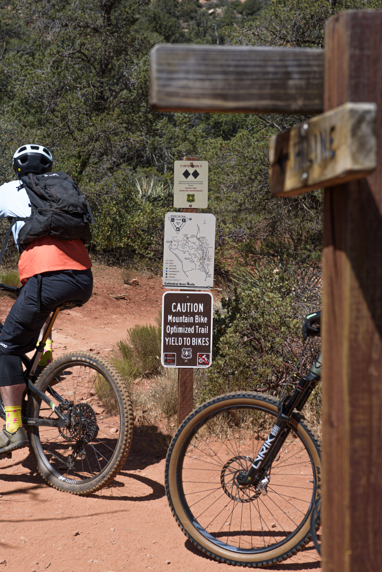 A mountain biker in a black backpack and orange shirt pauses on a dirt trail, with a wooden signpost nearby. The trail sign indicates that it is a mountain bike optimized trail, advising to yield to bikes. Lush vegetation surrounds the area, providing a natural backdrop.