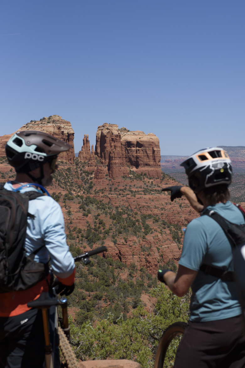 Two mountain bikers pause to admire a panoramic view of red rock formations under a clear blue sky. One biker, wearing a helmet, points toward the distant rock formations, while the other looks on with interest. The landscape features vibrant green vegetation contrasting with the reddish-brown terrain, capturing the beauty of the outdoor setting.