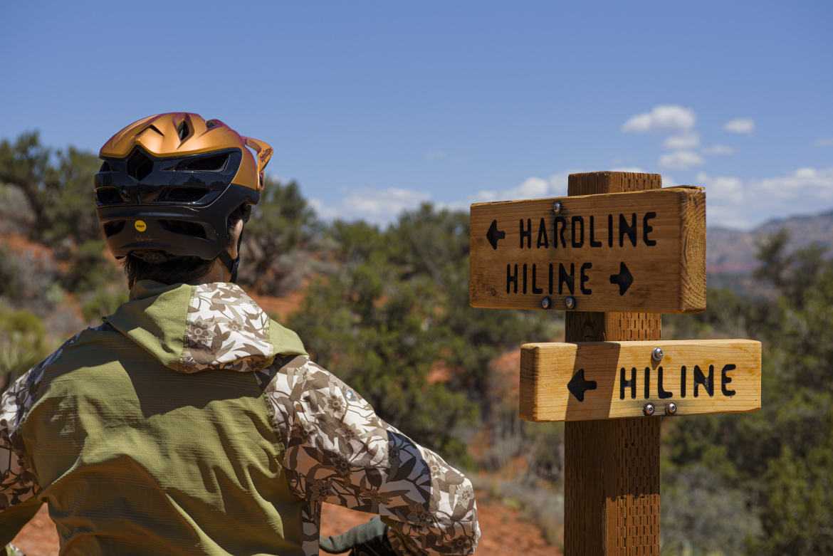 A person wearing a helmet and a patterned jacket stands facing a wooden trail sign with directional arrows indicating "Hardline" to the left and "Hiline" to the right. The background features a desert landscape with sparse vegetation under a clear blue sky.