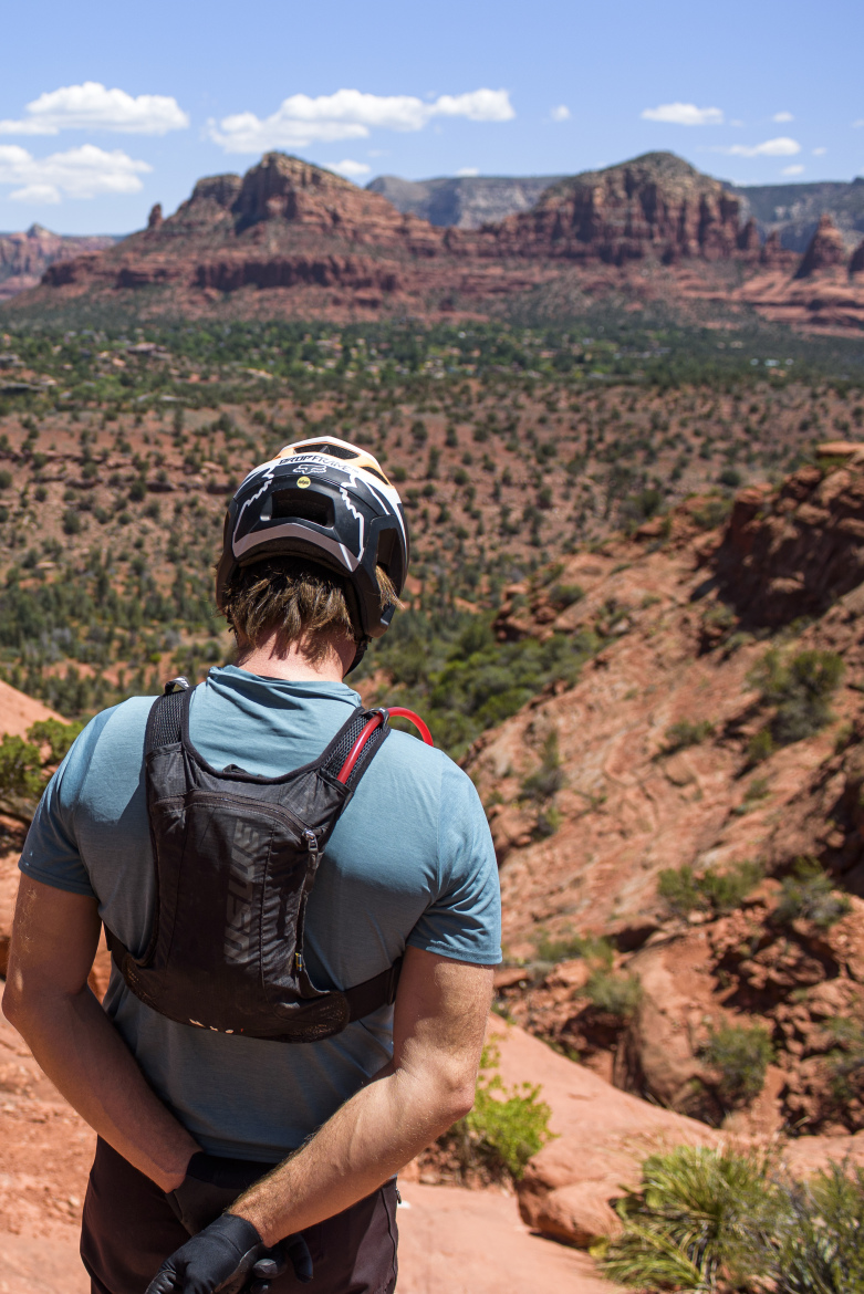 A person wearing a helmet and a backpack stands on a rocky outcrop, looking out over a vast landscape of red rock formations and sparse greenery under a bright blue sky with a few clouds.