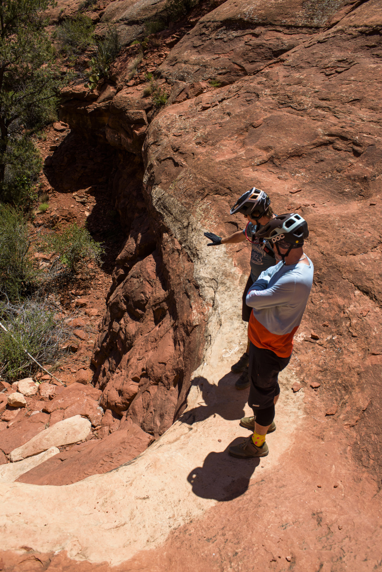 Two mountain bikers in protective gear stand on a rocky ridge, assessing a steep descent. The surrounding landscape features reddish-brown rocks and sparse vegetation typical of desert terrain. One rider gestures towards the trail ahead, while the other observes, both appearing to discuss the path they will take.