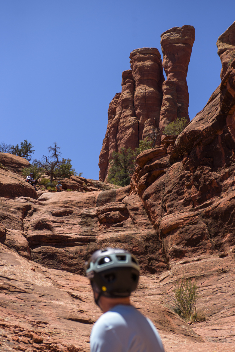 A person wearing a helmet looks up towards towering red rock formations under a clear blue sky, with other climbers visible on the rocky terrain in the background.
