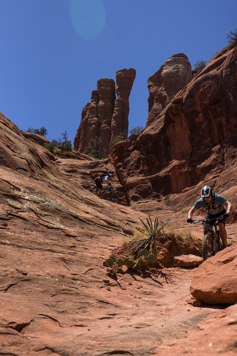 Two mountain bikers navigate a rocky trail in a desert landscape, with towering red rock formations in the background under a clear blue sky. The terrain features smooth and rugged surfaces, with some vegetation peeking through the rocks.