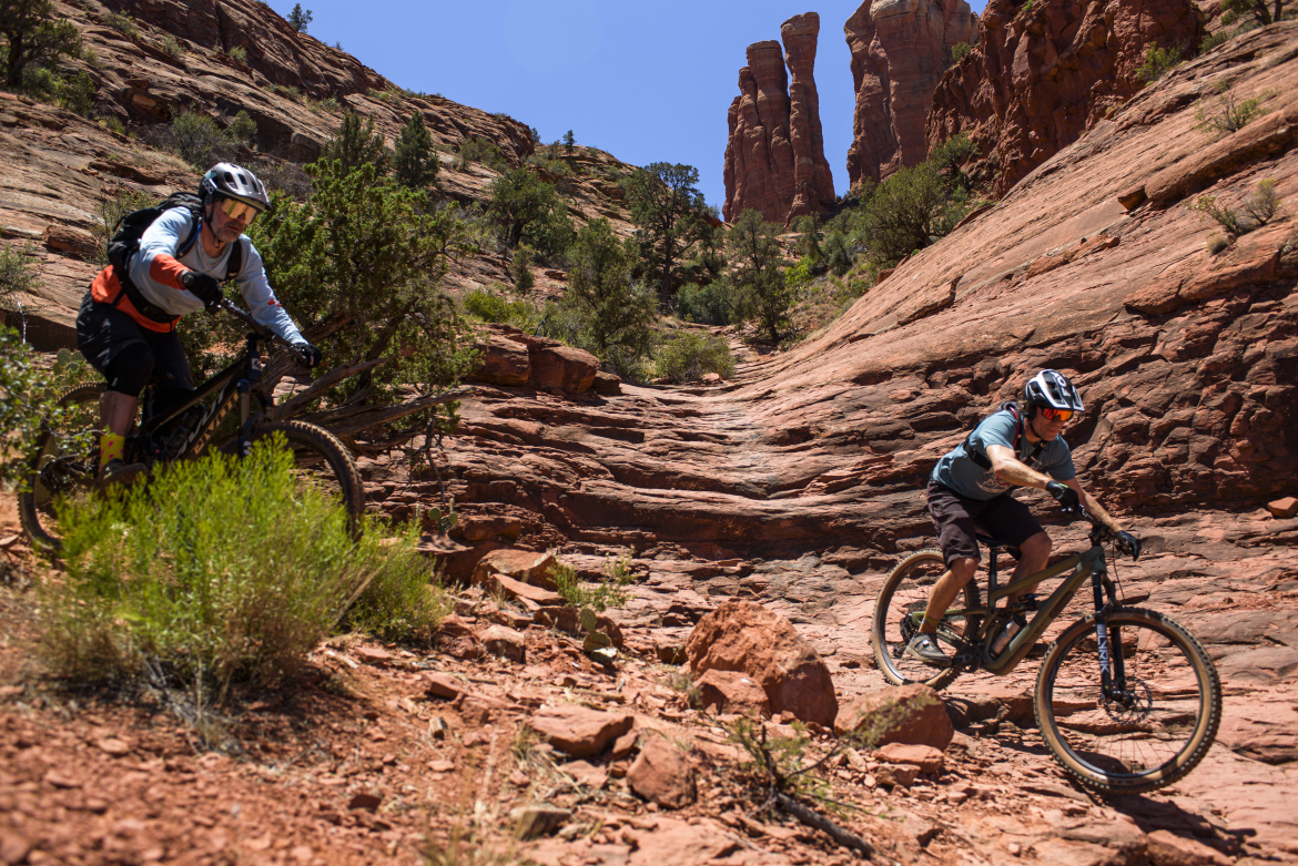 Two mountain bikers navigate a rocky trail surrounded by red rock formations and greenery under a clear blue sky. One rider is in the foreground, leaning forward as they maneuver over the terrain, while the other is slightly behind, also riding on the uneven path.