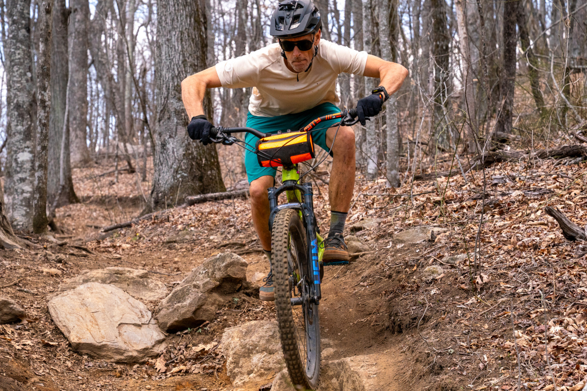 A mountain biker navigating a rocky trail in a forested area during early spring. The cyclist is wearing a helmet and sunglasses, crouched low over the handlebars. Surrounding trees appear bare, with a mix of dirt and fallen leaves covering the ground.