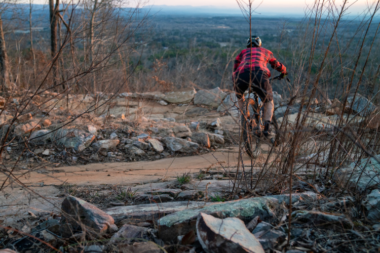 A mountain biker riding along a rocky trail at sunset, with a view of distant hills and trees in the background. The biker wears a black and red flannel shirt, and the foreground features scattered rocks and sparse vegetation.