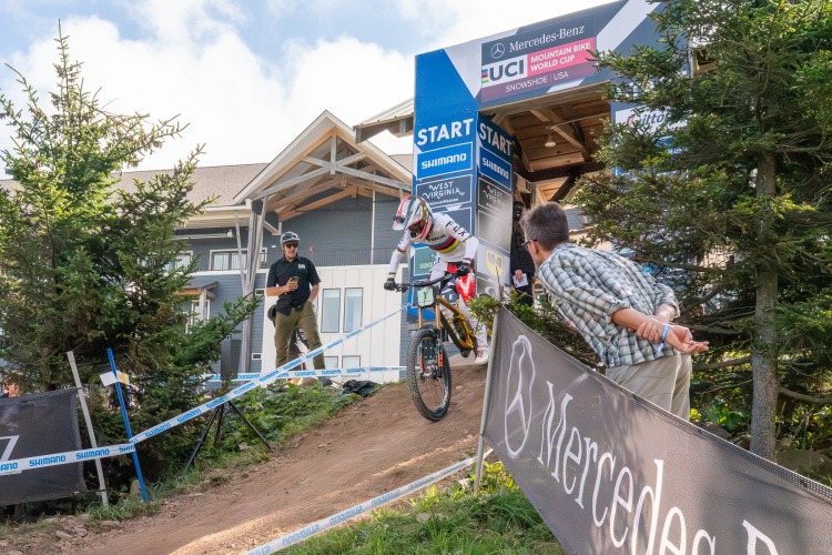 A mountain biker in a racing outfit prepares to start on a dirt track at the UCI Mountain Bike World Cup event. Two spectators watch from the sidelines, one holding a beverage. The setting features greenery and a building in the background, with event signage indicating the start of the race.