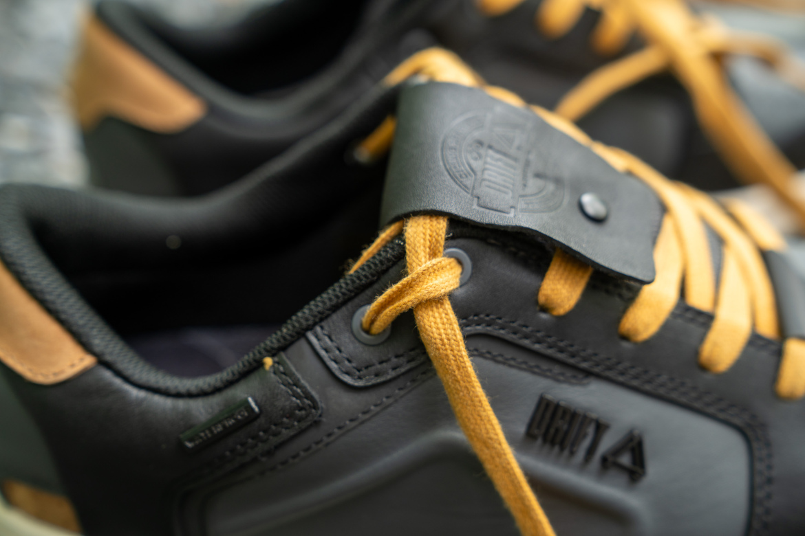 Close-up image of a pair of black sneakers featuring yellow laces and a black logo tag on the tongue. The design includes leather and fabric materials with a contrasting tan detail on the heel.