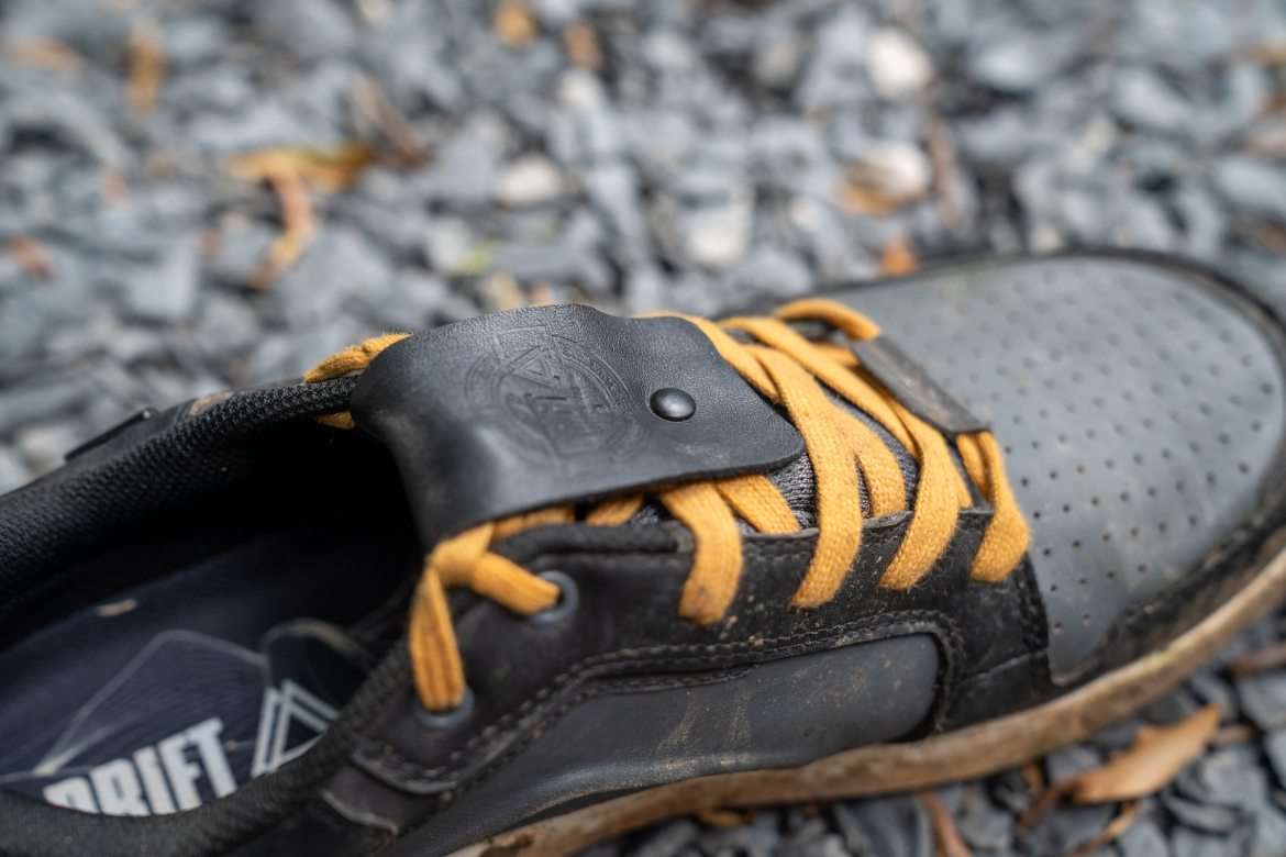 Close-up of a black sneaker with yellow laces, featuring a leather strap with a button closure. The shoe is partially dirty, set against a background of gray gravel and scattered leaves.