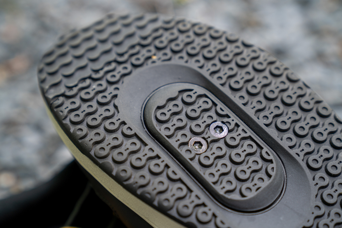 Close-up view of the textured sole of a shoe, featuring a unique tread pattern with small dots and waves, along with two metallic cleats positioned in the center. The background is blurred, emphasizing the shoe's details.