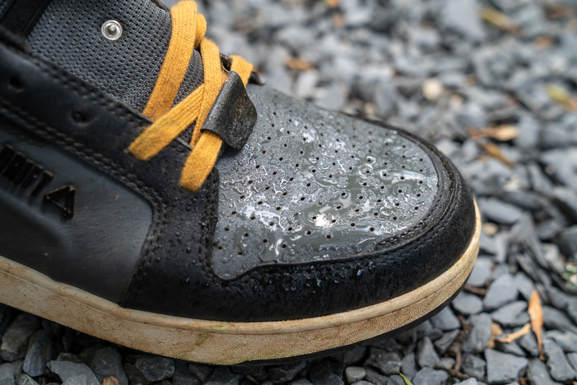Close-up of a black sneaker with yellow laces, showing water droplets on the surface, resting on a bed of small gray stones.